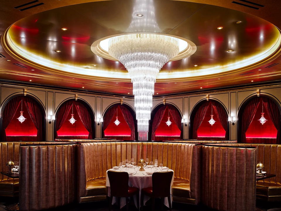 A glamorous dining room with gold leather booths, a tiered crystal chandelier, and arched alcoves with red velvet curtains.