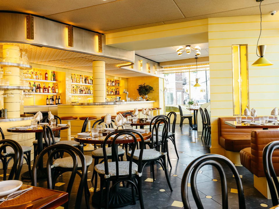 A bright yellow-toned dining room at San Sabino, featuring a curved bar with stools, multiple round tables with bentwood chairs, and brown leather