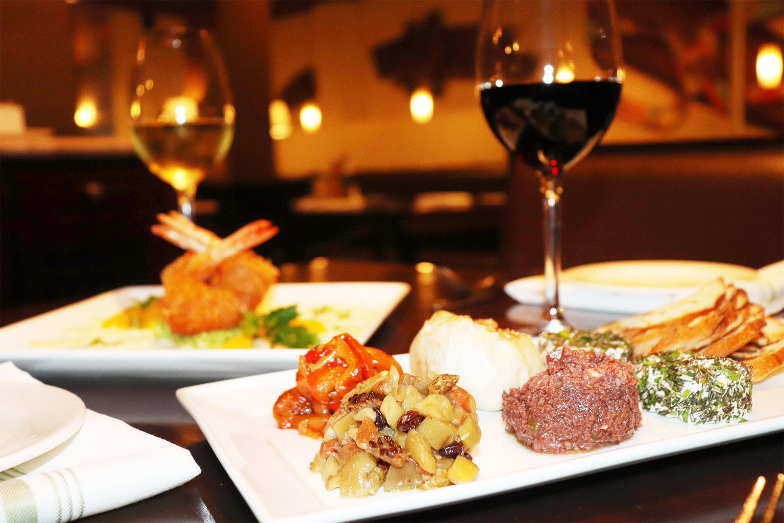 A white plate with various appetizers, including fried shrimp, a spread, and bread, alongside two wine glasses in a restaurant.
