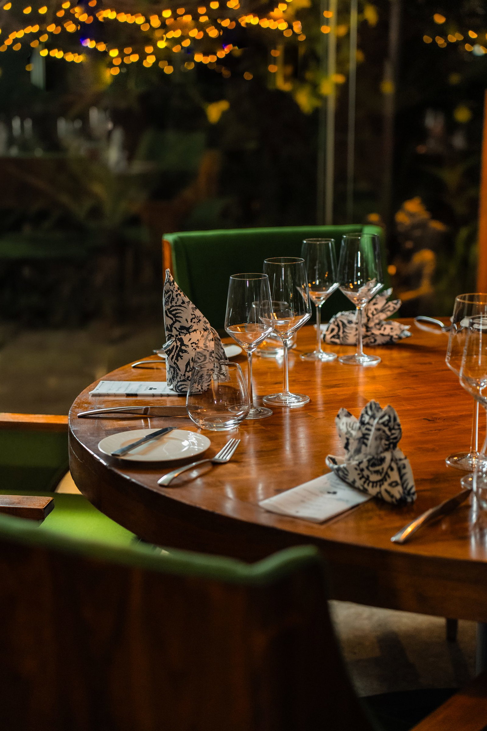 A round wooden dining table is set for multiple guests with wine glasses, plates, and patterned napkins.