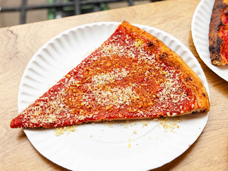 A triangular slice of Roman-style pizza from Fini Pizza, topped with tomato sauce and grated cheese, on a white paper plate.