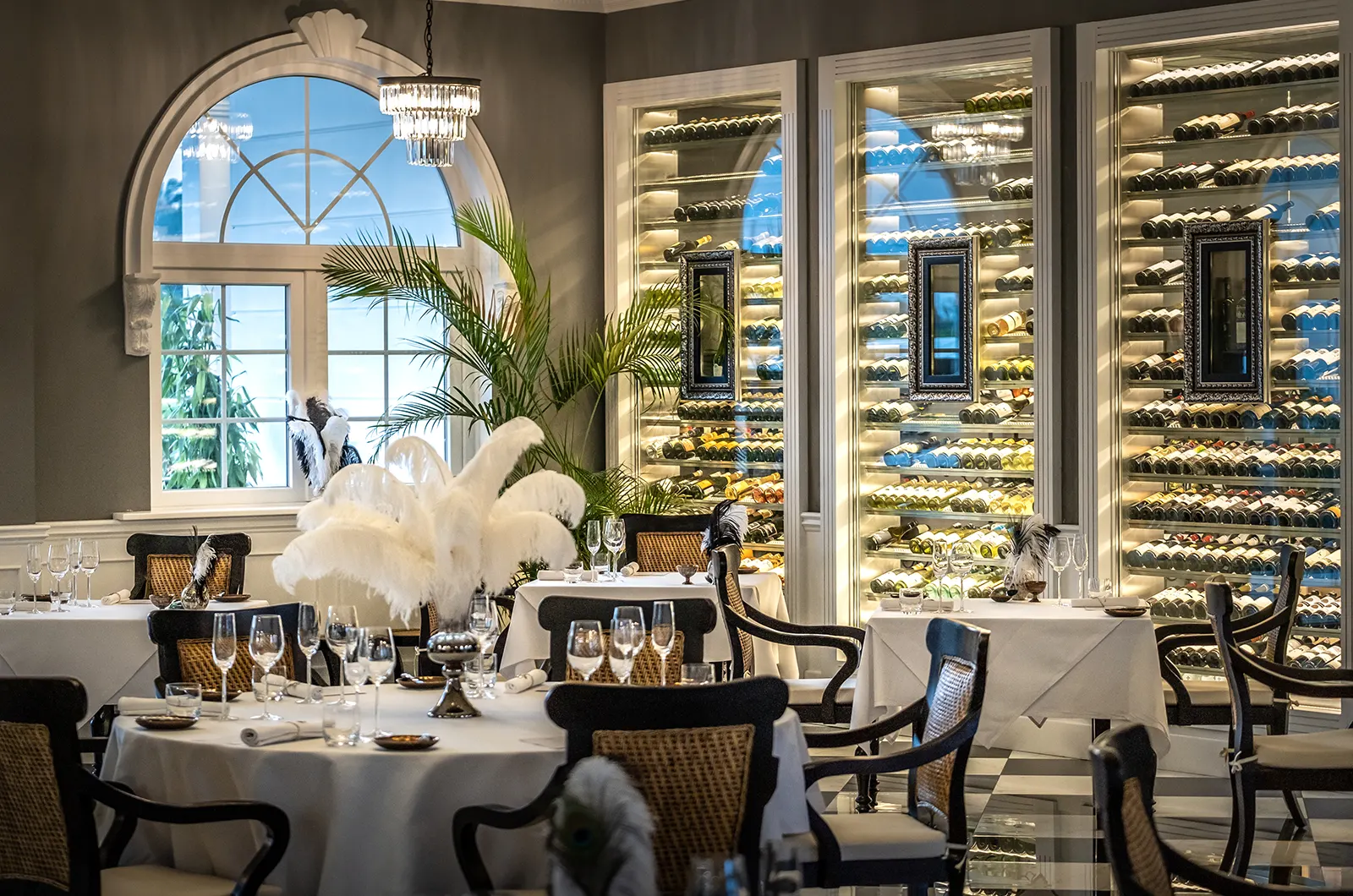 A fine dining restaurant with white tablecloths, wicker chairs, and a large arched window. Three tall wine cellars with illuminated bottles line one
