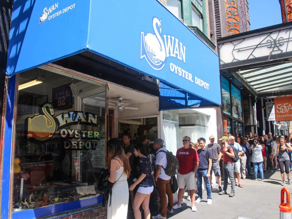 A long line of people queues outside Swan Oyster Depot, a seafood restaurant with a blue awning and a swan logo.