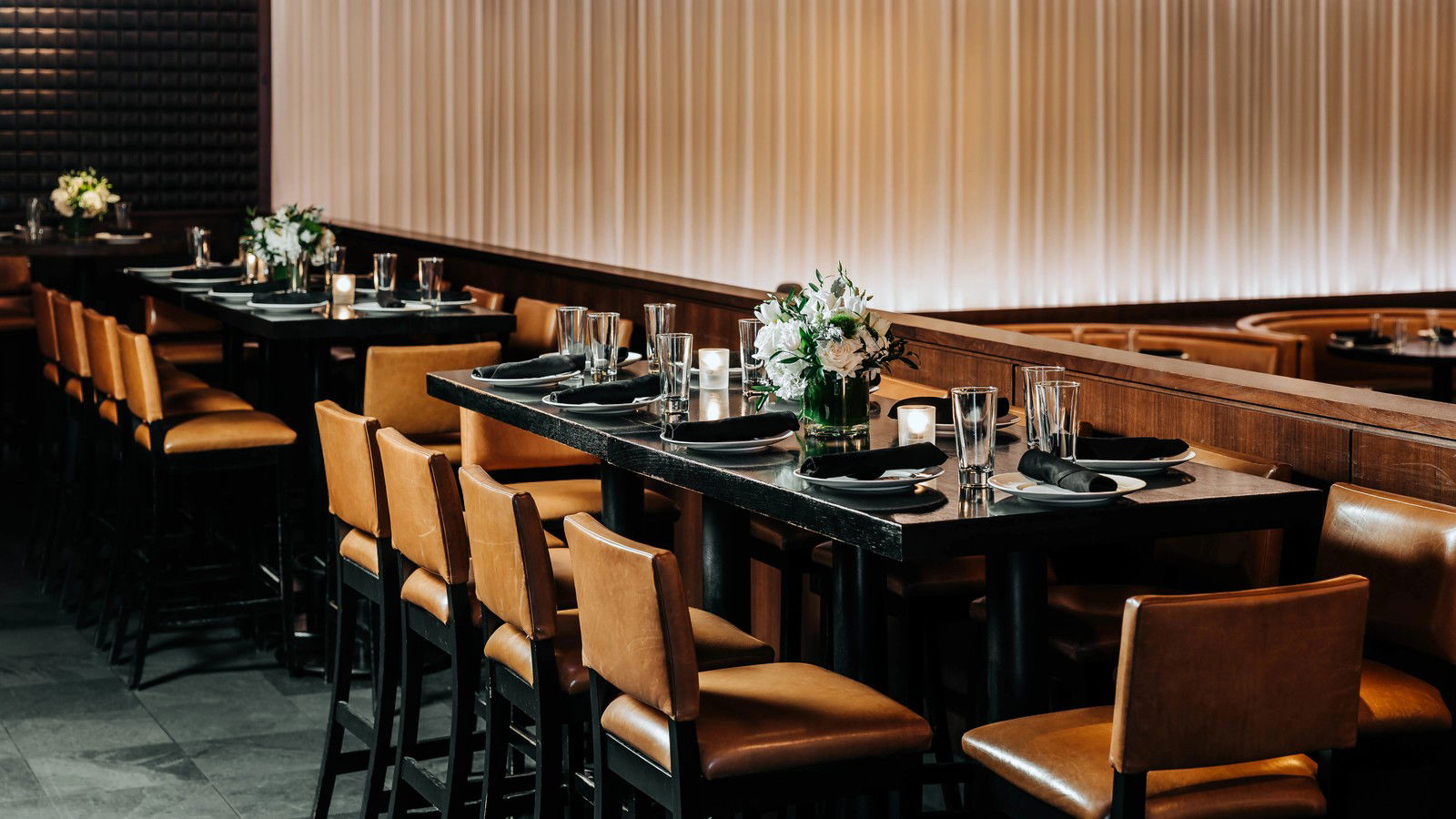 An upscale steakhouse dining room with high-top tables, leather chairs, floral centerpieces, and a glowing ribbed wall.