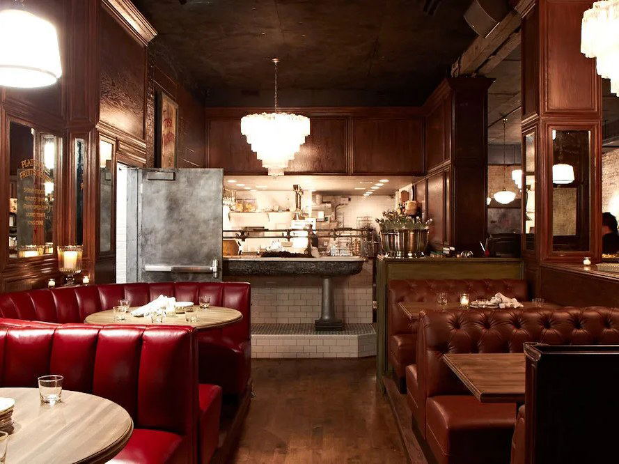 The interior of a dimly lit restaurant with red leather banquettes, brown tufted leather booths, dark wood paneling, and an open kitchen.