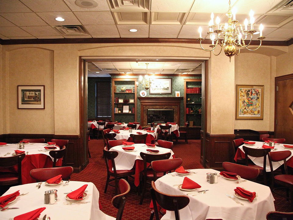 A classic dining room with white tablecloths, red napkins, dark wood chairs, red carpet, a chandelier, and a fireplace.