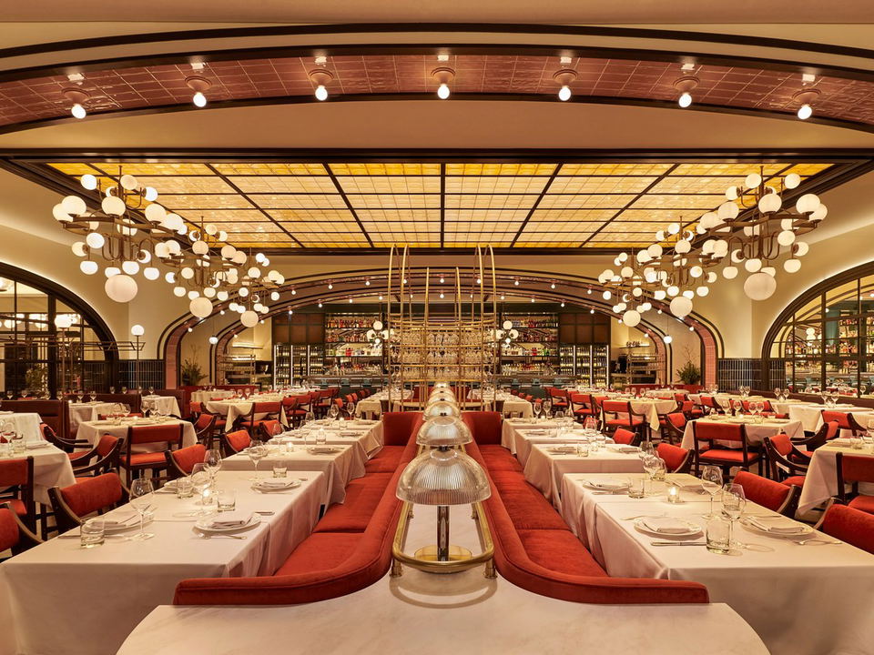 A grand, Art Deco-style restaurant dining room with red velvet banquette seating, white-linen tables, globe pendant chandeliers, a glowing amber