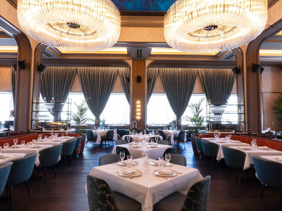 An elegant dining room with two large crystal chandeliers, teal chairs, white-tablecloth tables, and a blue ceiling.
