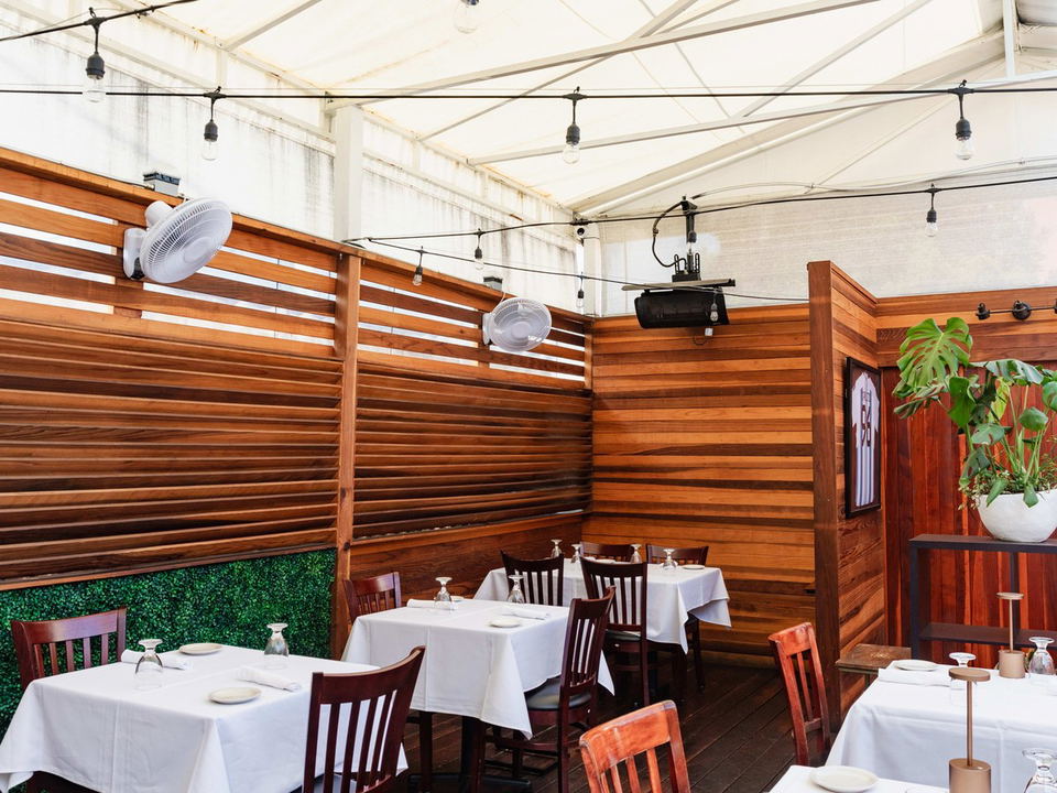 A covered outdoor patio dining room with warm wood-slat walls, white-tablecloth tables, string lights, wall-mounted fans, and tropical plant décor.