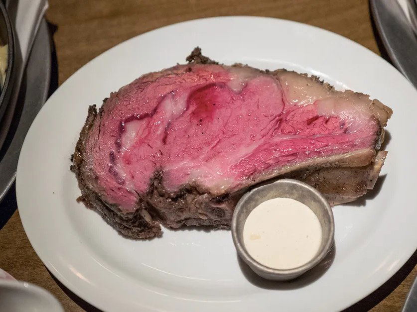 A close-up of a thick, bone-in prime rib slice on a white plate with a small ramekin of horseradish sauce.