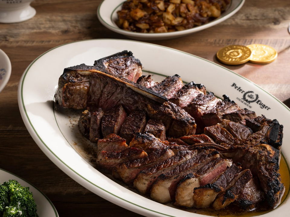 A sliced porterhouse steak on a white oval platter with a green rim, branded with "Peter Luger Steak House," accompanied by hash browns and broccoli.