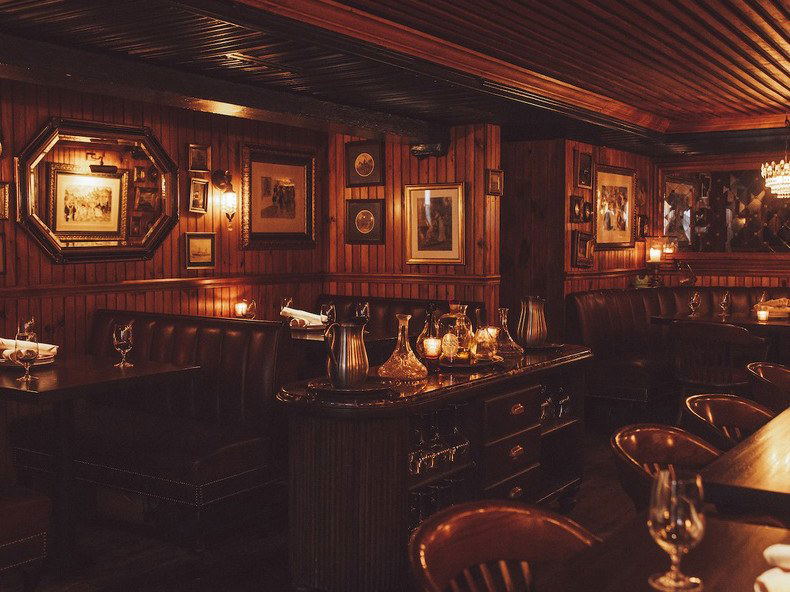 A dimly lit, wood-paneled dining room with dark leather banquettes, framed artwork, and a central service station.