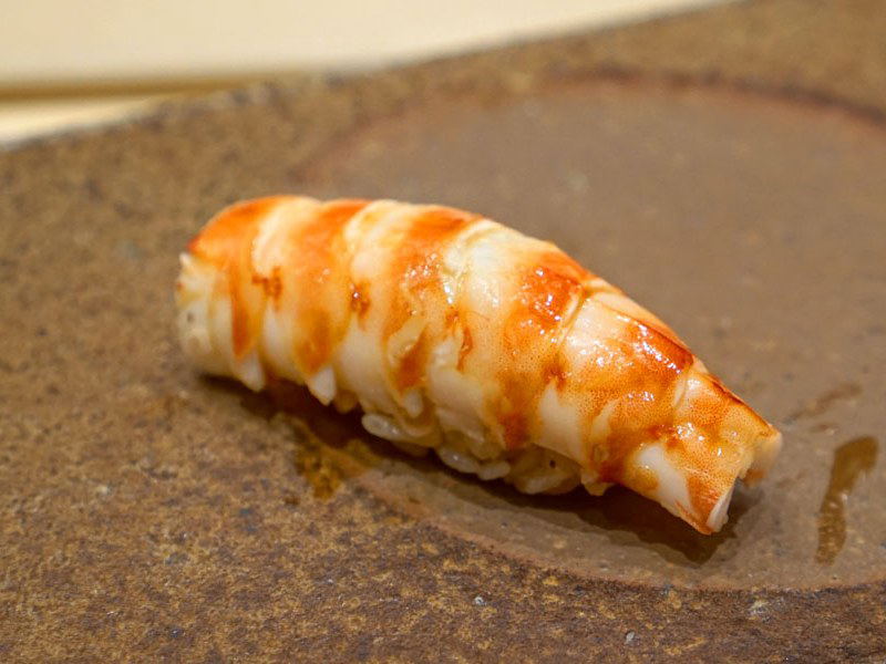 A single piece of nigiri sushi, identified as Kasugo Dai, with a reddish-orange seared top and white rice base, rests on a rustic brown ceramic plate.