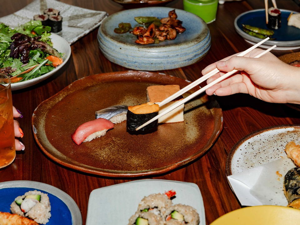 A hand uses chopsticks to pick up a piece of uni gunkan sushi from a rustic brown plate, surrounded by other Japanese dishes.