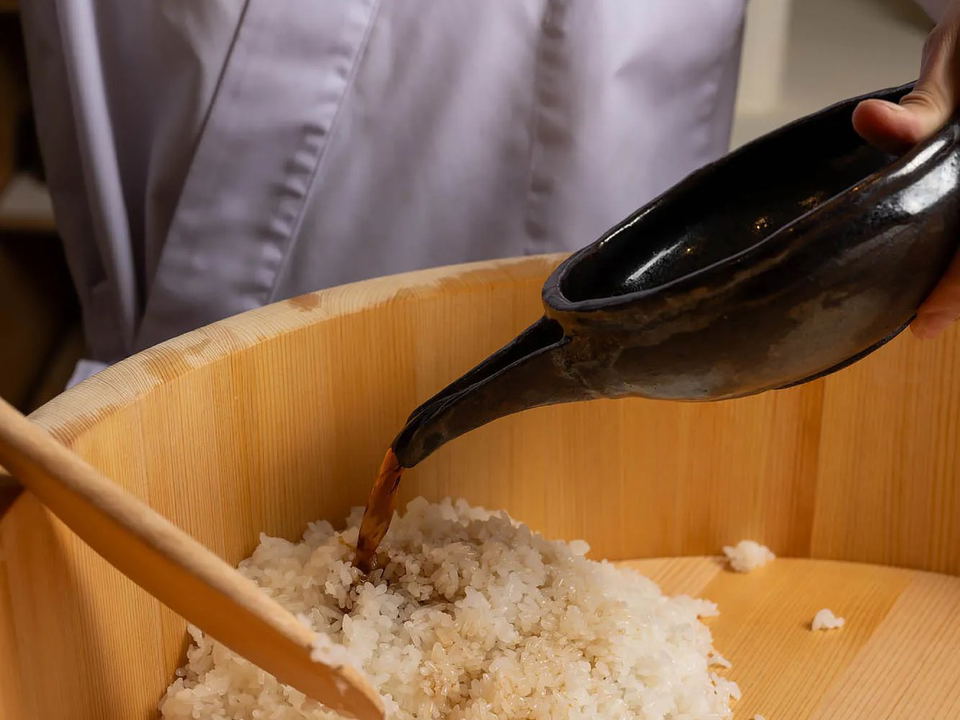 A chef in a white coat pours seasoned vinegar from a dark ceramic vessel onto cooked rice in a wooden hangiri (sushi rice tub), with wooden rice.