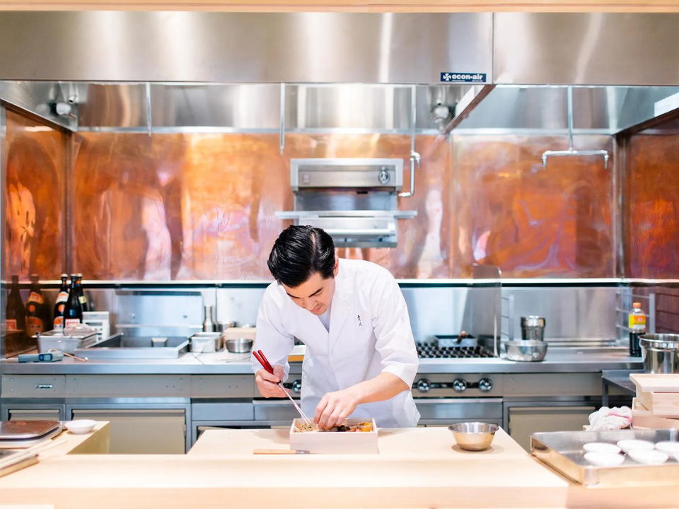 Chef Brandon Go, in a white coat, meticulously plates a dish with red chopsticks at a light wood counter in an open kitchen featuring copper walls