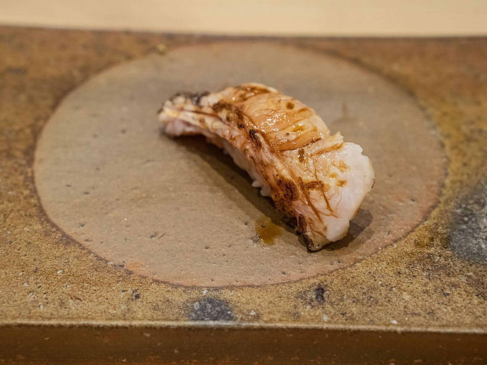 A single piece of lightly seared nigiri sushi, with a pale fish topping showing grill marks, rests on a rustic, round ceramic plate.