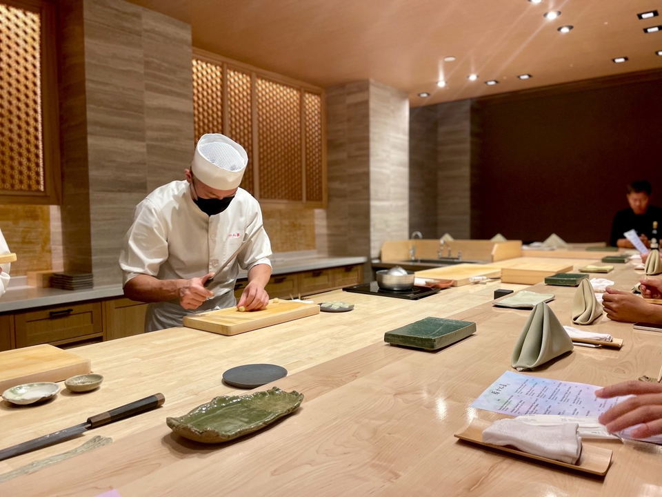 A chef in a white uniform and black mask meticulously slices ingredients on a wooden cutting board at a light wood omakase counter in Sushi Kaneyoshi.