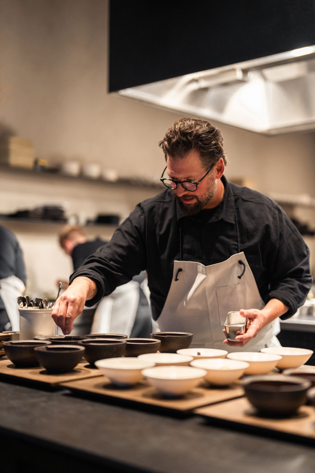 A chef in black chef's coat and glasses plating dishes in a modern kitchen