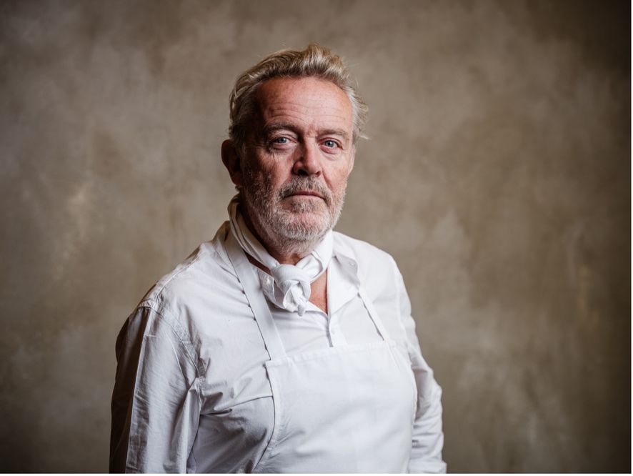 Professional portrait of a middle-aged man with gray beard wearing white chef's attire against a neutral background.