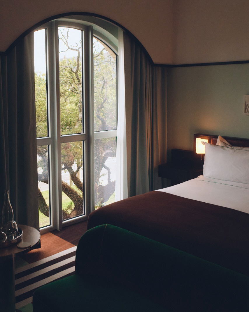 A hotel guest room with arched doorway, large windows overlooking trees, green velvet bedding, warm wood flooring with striped rug, and 1920s-inspired
