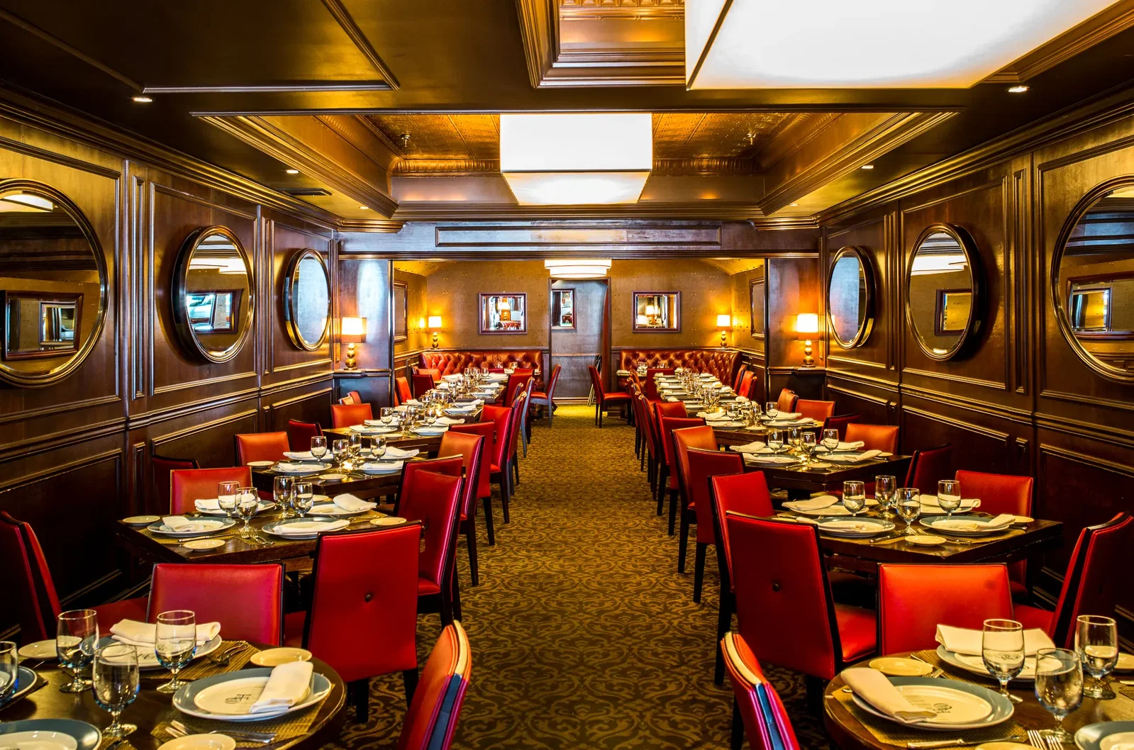 A long, elegant dining room with dark wood paneling, round mirrors, and tables set with white tablecloths and red chairs.