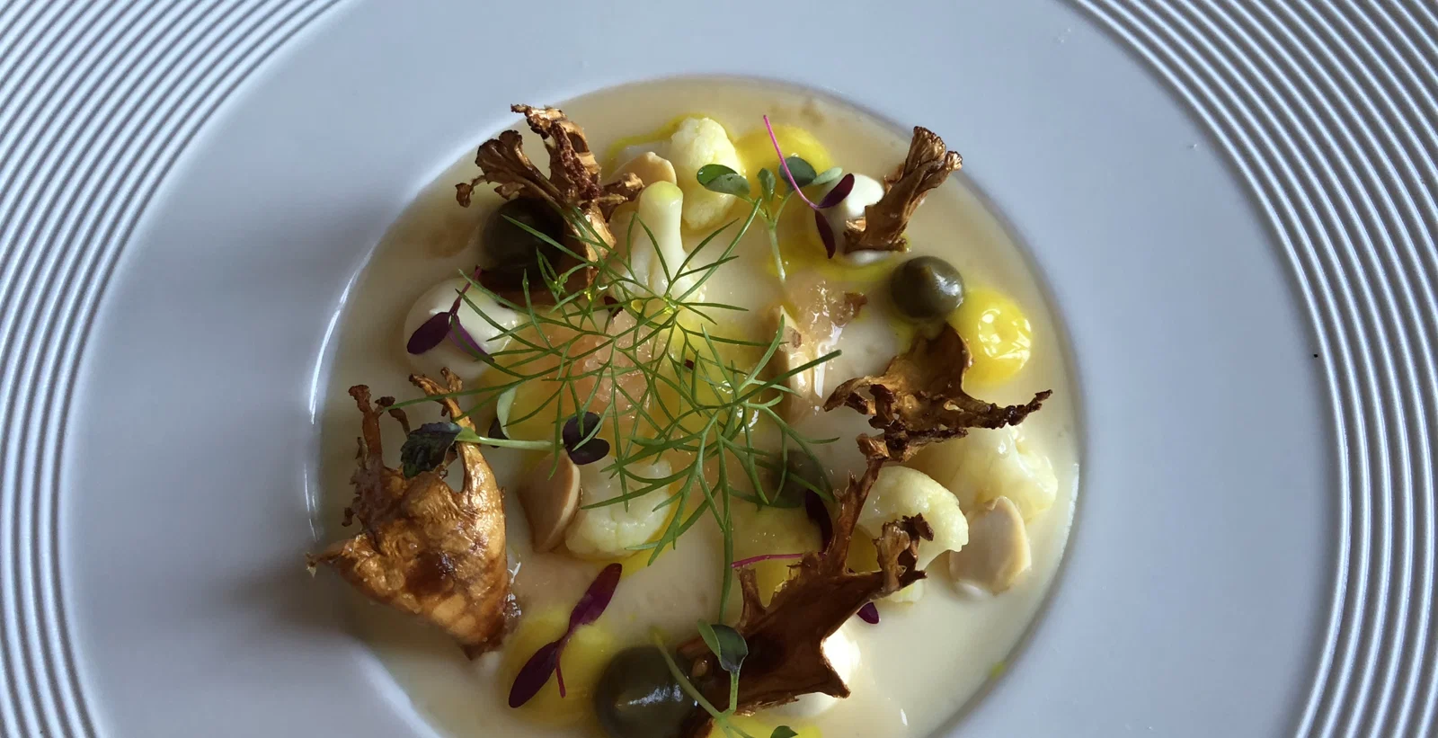 An overhead shot of a fine dining dish, featuring white cauliflower florets, brown crispy mushrooms, green olives, and delicate herbs in a creamy.