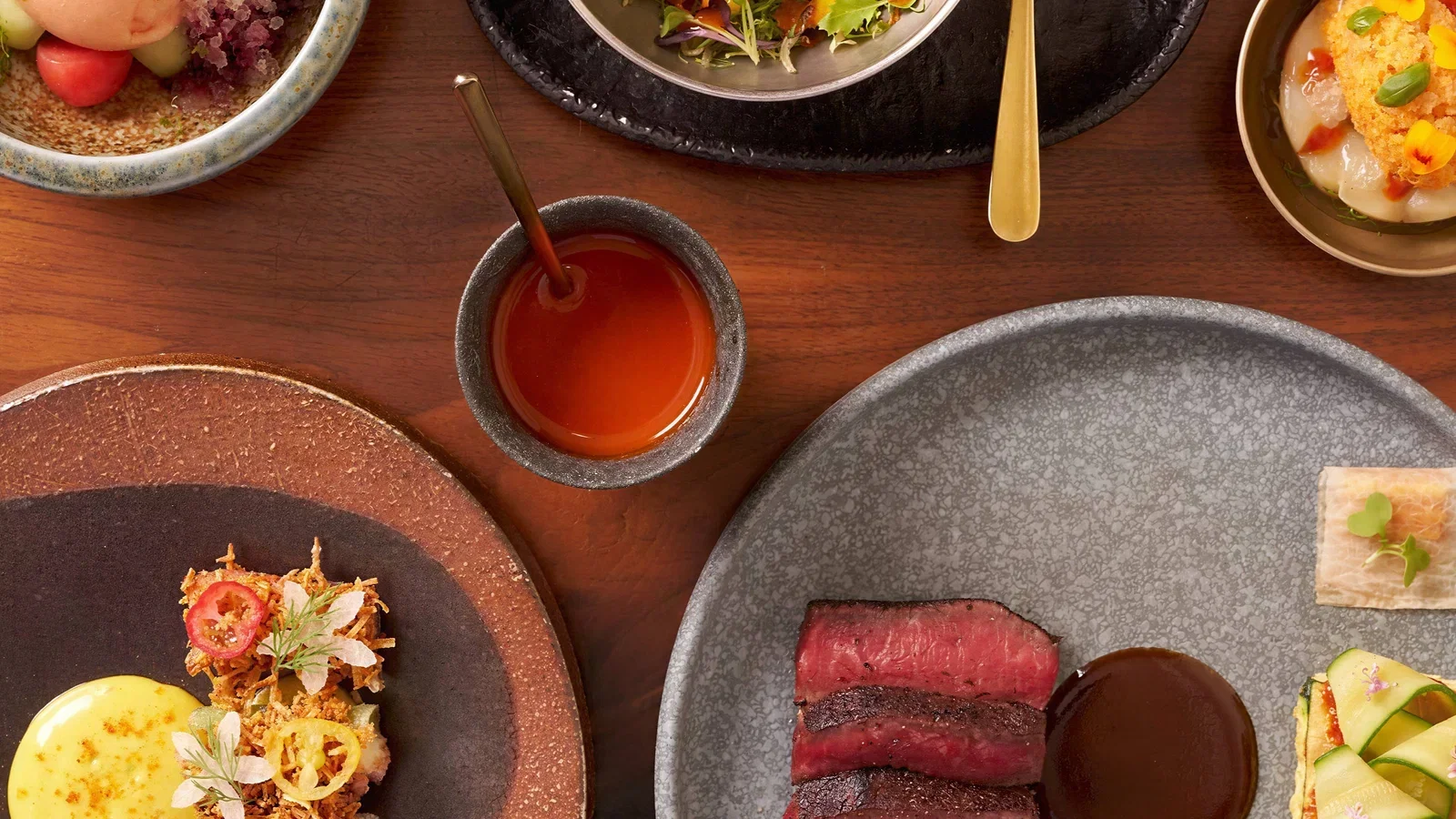 An overhead view of a table set with various dishes, including sliced steak, a bowl of red sauce, and other appetizers.