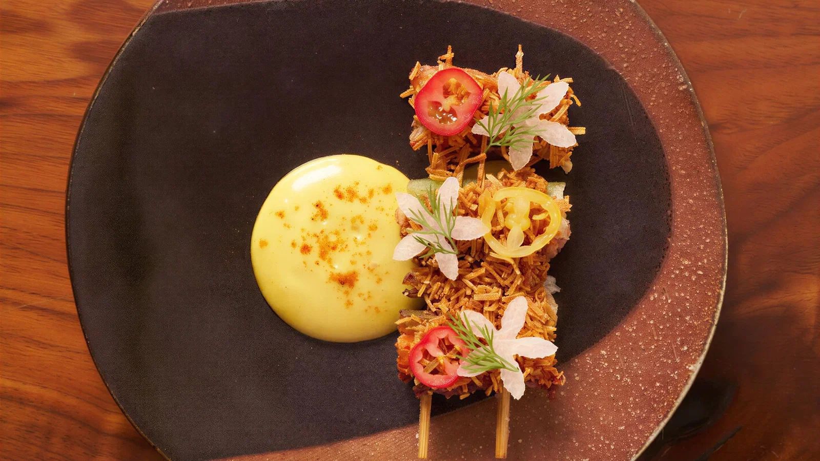 Two skewers of fried, crispy food with white flowers and red and yellow pepper slices, next to a dollop of yellow sauce on a dark plate.