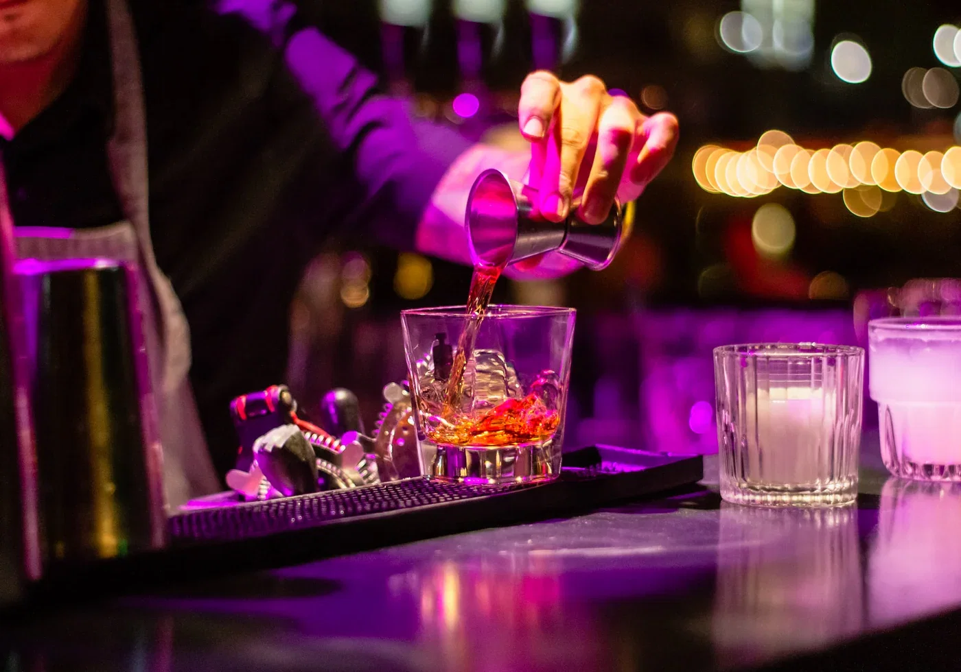 A bartender in a dimly lit bar pours a drink from a jigger into a glass with ice, illuminated by purple light.