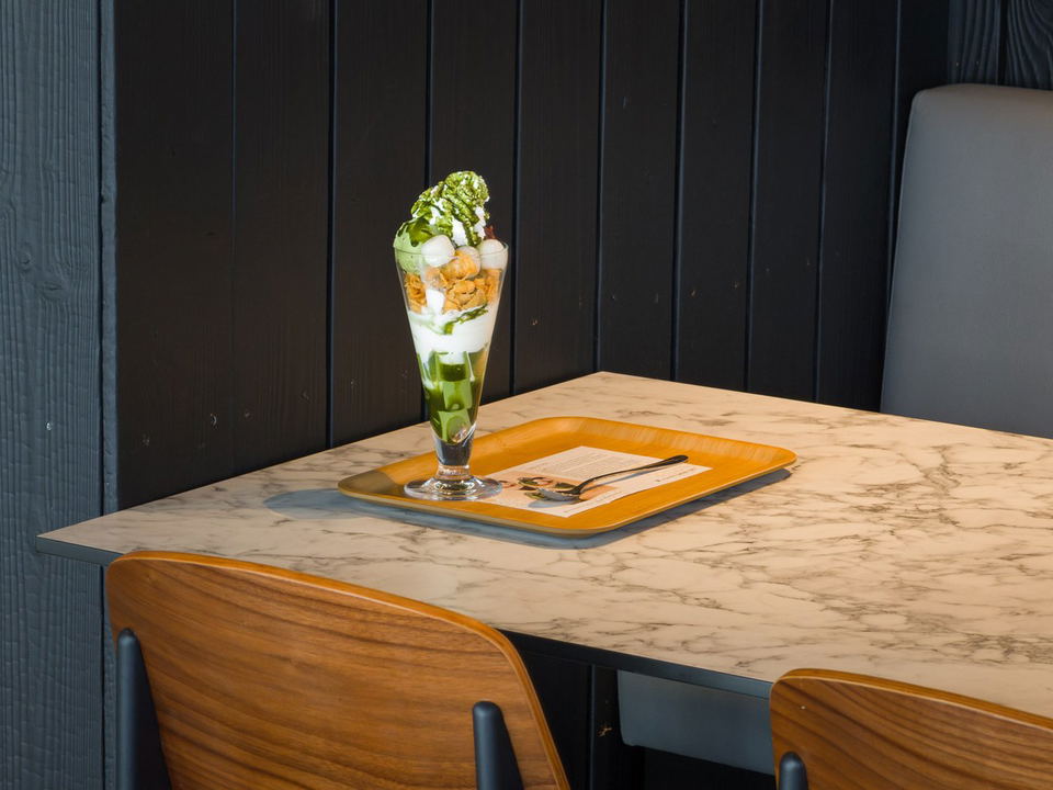A tall glass of matcha parfait dessert with green tea jelly, white cream, and various toppings sits on a wooden tray on a marble table.