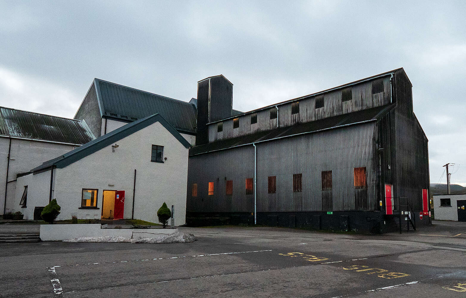 The exterior of the Tomatin Distillery in the Scottish Highlands, featuring a white building and a large corrugated metal structure under a cloudy sky.