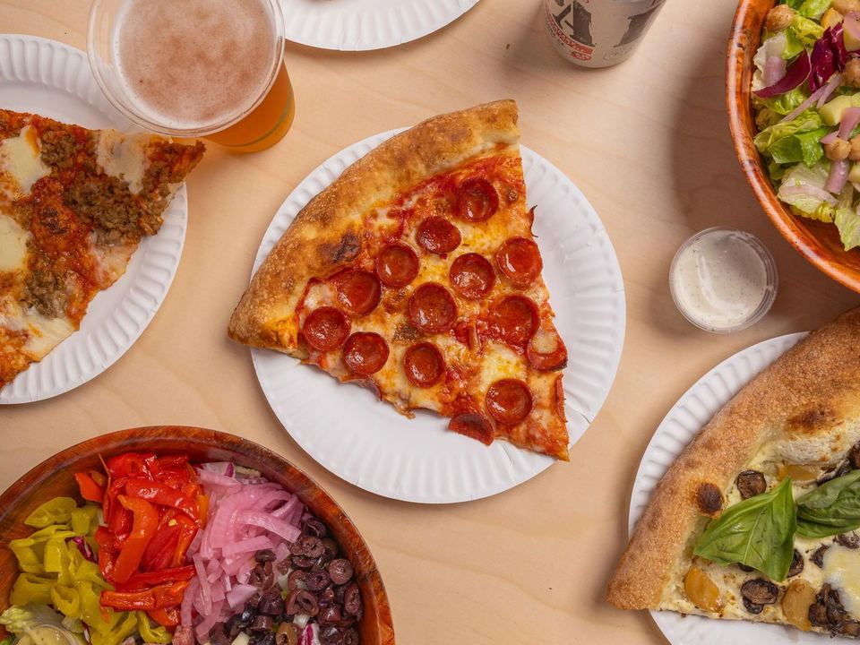 An overhead shot of various pizza slices, salad bowls, chicken wings, and beverages on paper plates and wooden bowls, showing a casual dining spread.