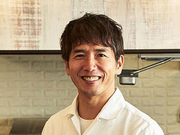 A smiling Chef Seizo Mitani, dressed in a white chef's coat with an embroidered sleeve, stands in a modern kitchen with wooden and tiled walls,
