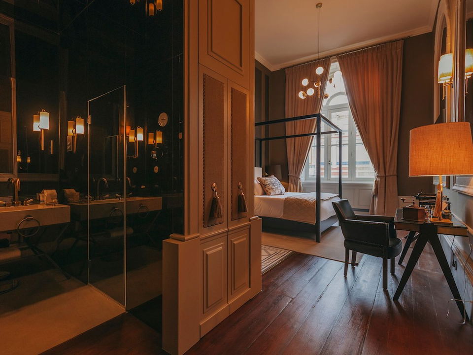 An upscale hotel bedroom with a black metal canopy bed, wooden vanity with mirror, desk area, orange curtains, and large windows with city views.