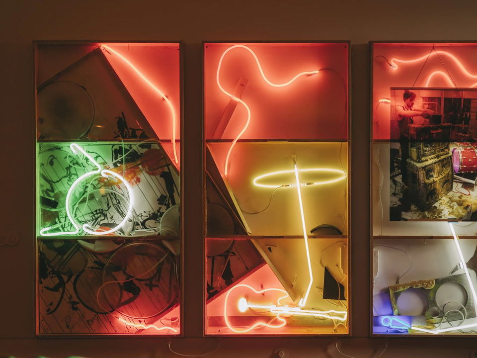 Three framed display boxes containing neon light artworks in red, green, yellow, and blue, with photographs and objects arranged behind the neon tubes