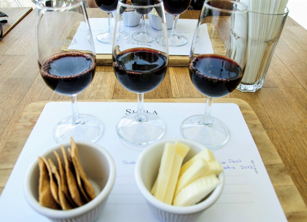 Wine tasting setup with three glasses of red wine on a white tray marked 'SUVLA', accompanied by crackers and cheese slices