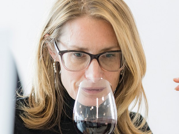 A woman with blonde hair and dark-framed glasses sniffs a glass of red wine at a tasting table surrounded by multiple wine glasses.