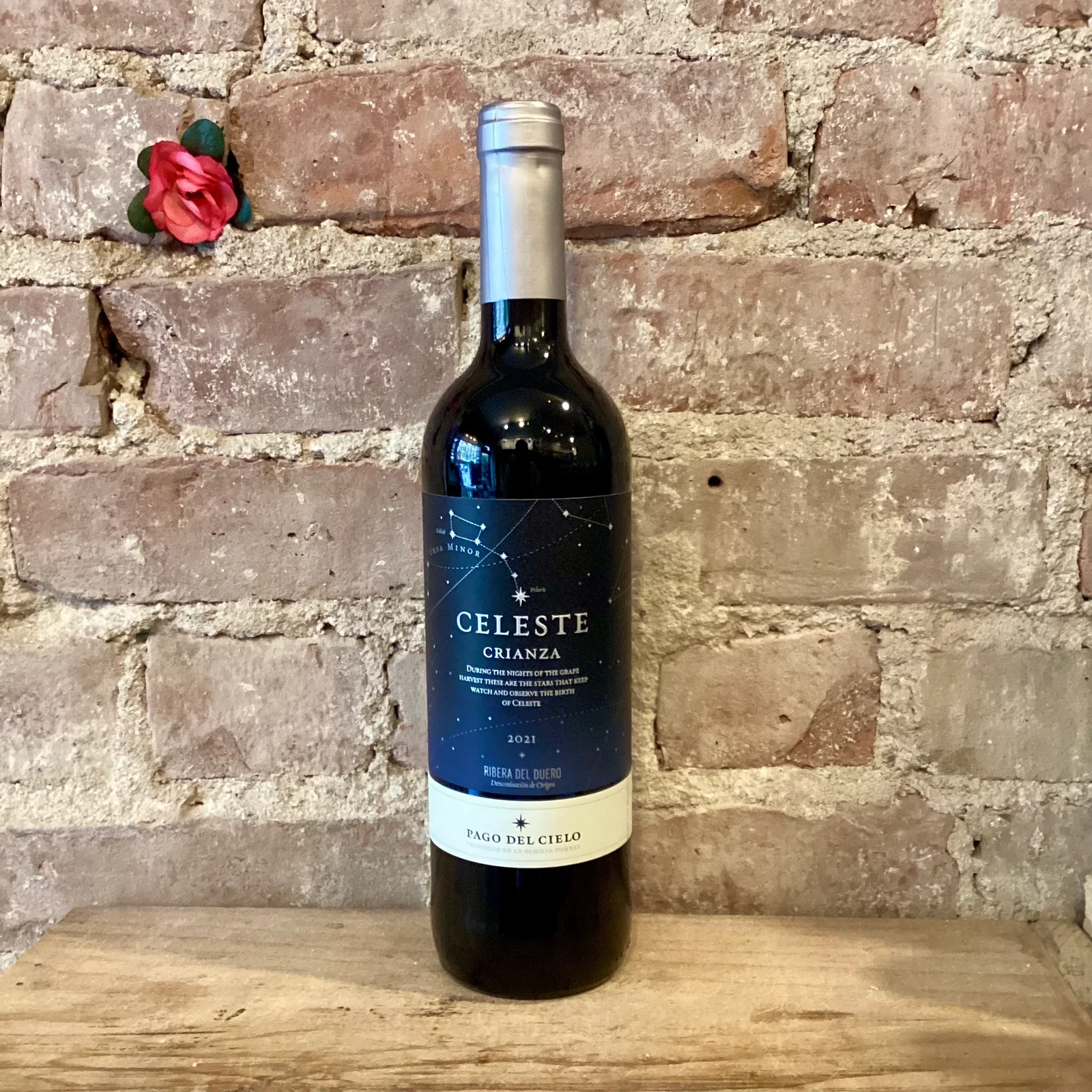 A dark bottle with a starry label and silver top rests on wood against a rustic brick wall, accented by a small red rose.