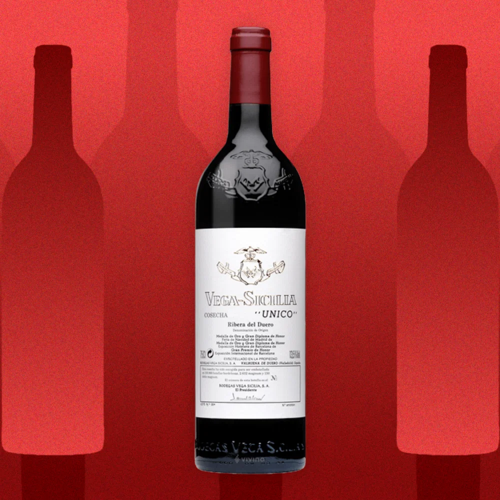 A dark bottle with a white label and red foil stands centrally against a textured red background, flanked by two subtle silhouettes.