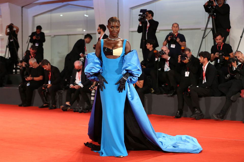 A woman in a striking blue evening gown with voluminous ruffled shoulders walks on a red carpet surrounded by photographers and media personnel in formal attire.