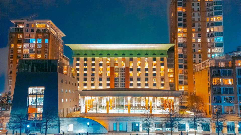 A modern hotel building with a green roof and many illuminated windows at night, flanked by taller residential buildings with glowing windows.