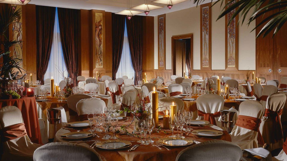 A formal dining room at Canary Riverside Plaza hotel, featuring multiple round tables set with white and gold place settings, candles, and floral