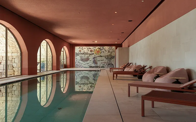 An indoor swimming pool with terracotta-colored arched ceiling and walls, stone arches on the left, a large mosaic artwork on the far wall, and lounge