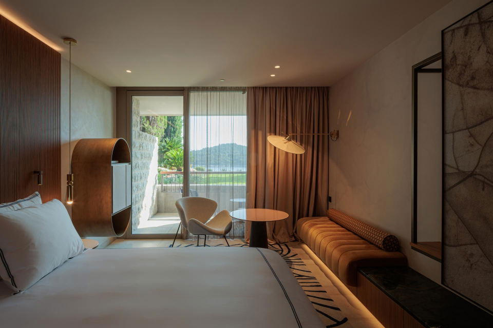 Luxury hotel bedroom with white bed, brown accent chair, round table, curved sofa, and glass doors opening to a scenic balcony view