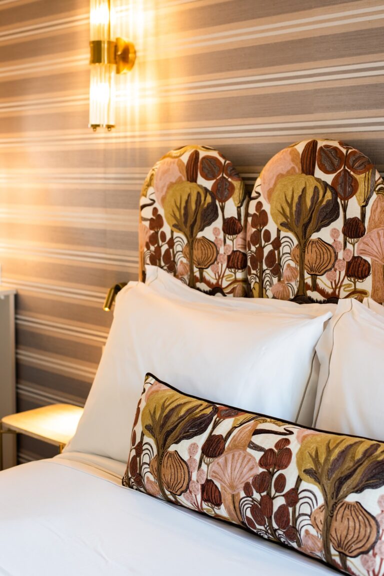 A hotel bedroom interior featuring a distinctive upholstered headboard with botanical print fabric in browns, pinks, and golds, white bedding, and str