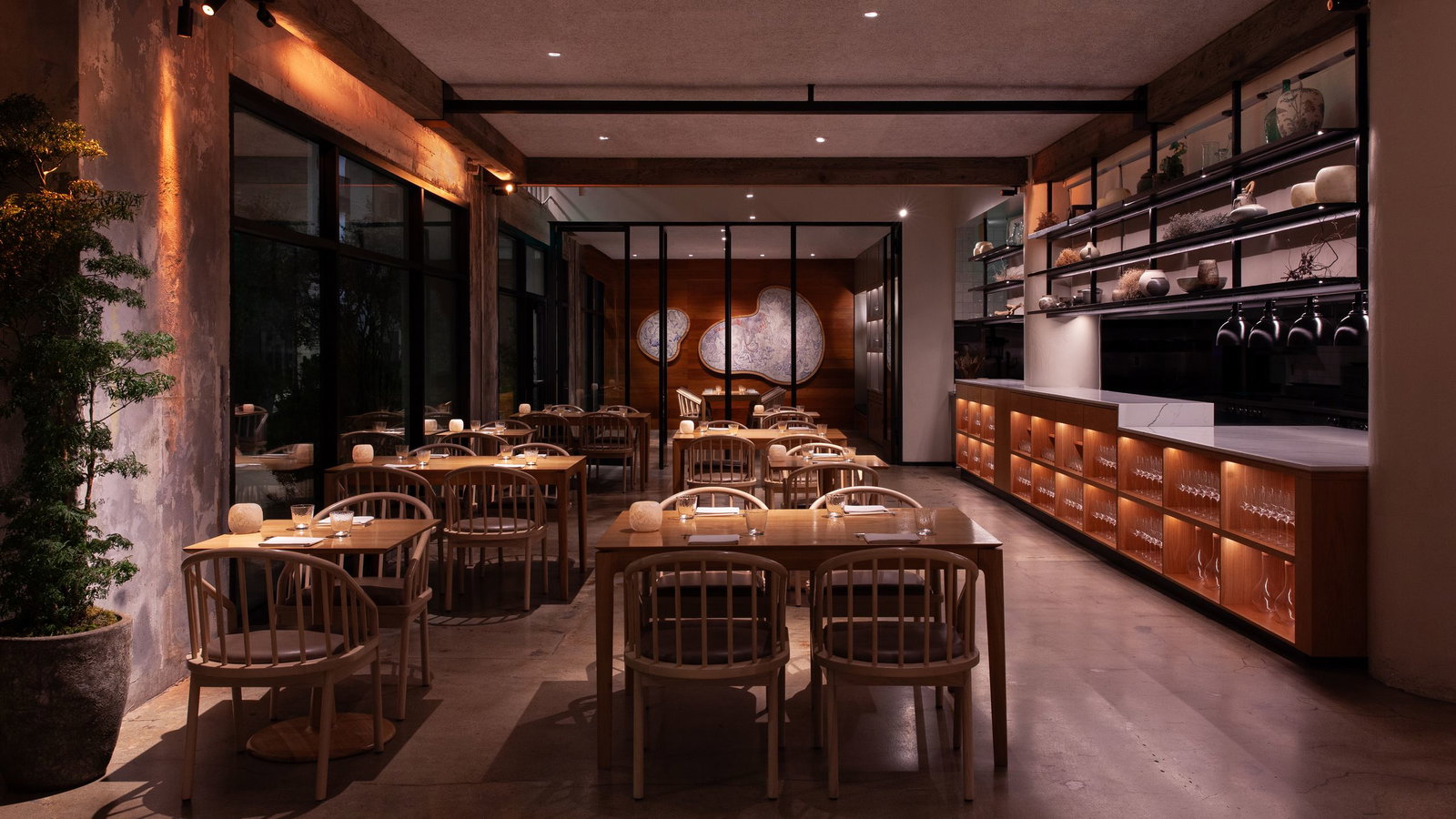 A warmly lit, intimate restaurant dining room with light wood chairs and tables, open shelving with glassware, and large glass windows.