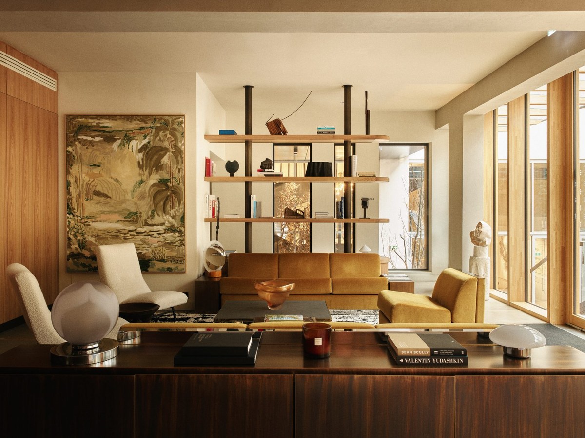 Modern living room with wooden shelving unit, tan sofa, cream accent chair, dark coffee table, and large windows with natural light.