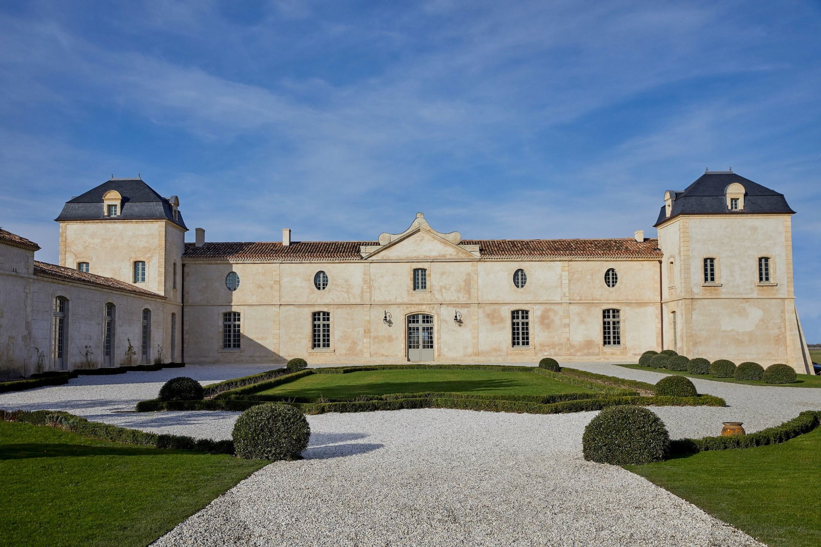 Exterior view of a large cream-colored château with two prominent towers, black mansard roofs, multiple tall windows, and formal gardens with boxwood hedges and gravel walkways under a blue sky.