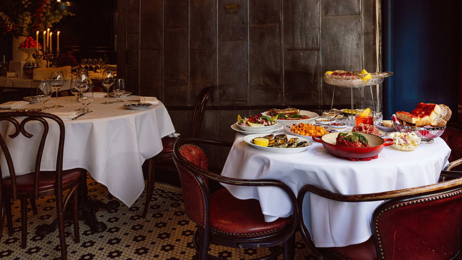 A dining room at Carbone New York with white tablecloths, red leather chairs, dark paneled walls, and a spread of Italian-American dishes.